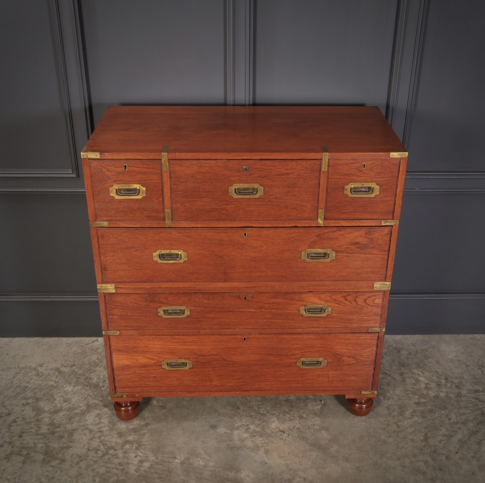 A Victorian teak military campaign secretaire chest featuring an open fall-front desk compartment, flush brass handles, brass corner brackets, and turned feet against a dark grey panelled wall. View II