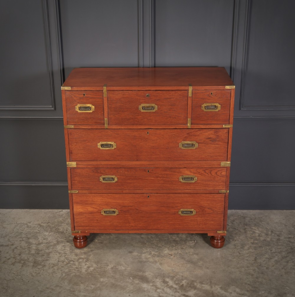 A Victorian teak military campaign secretaire chest featuring an open fall-front desk compartment, flush brass handles, brass corner brackets, and turned feet against a dark grey panelled wall. View III