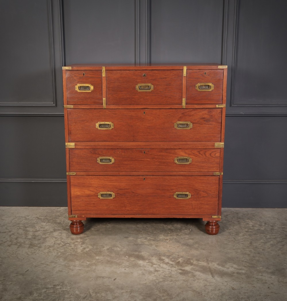 A Victorian teak military campaign secretaire chest featuring an open fall-front desk compartment, flush brass handles, brass corner brackets, and turned feet against a dark grey panelled wall. View V