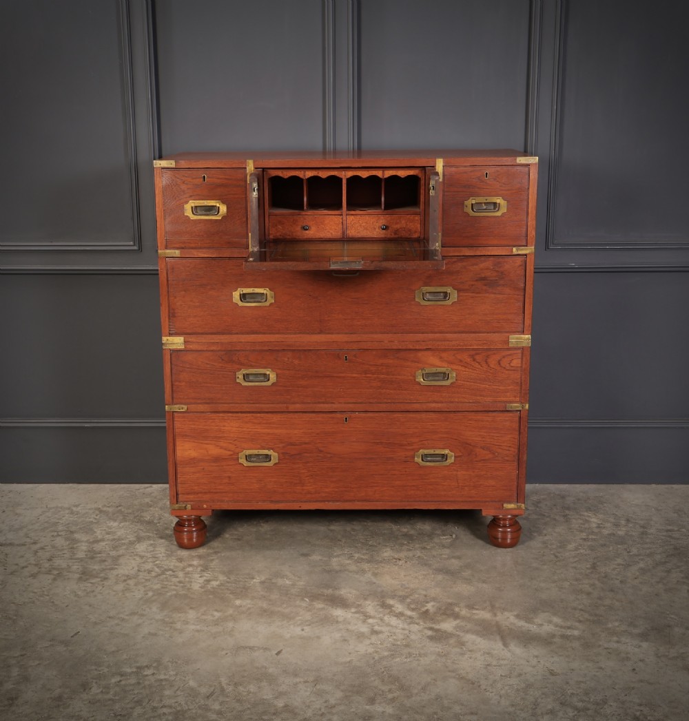 A Victorian teak military campaign secretaire chest featuring an open fall-front desk compartment, flush brass handles, brass corner brackets, and turned feet against a dark grey panelled wall.