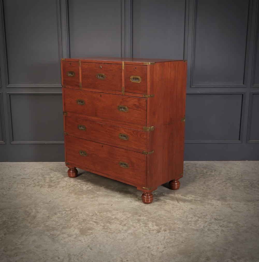 A Victorian teak military campaign secretaire chest featuring an open fall-front desk compartment, flush brass handles, brass corner brackets, and turned feet against a dark grey panelled wall. View VIII