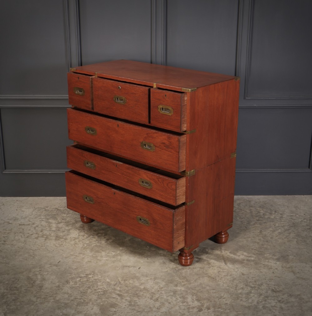 A Victorian teak military campaign secretaire chest featuring an open fall-front desk compartment, flush brass handles, brass corner brackets, and turned feet against a dark grey panelled wall. View IX