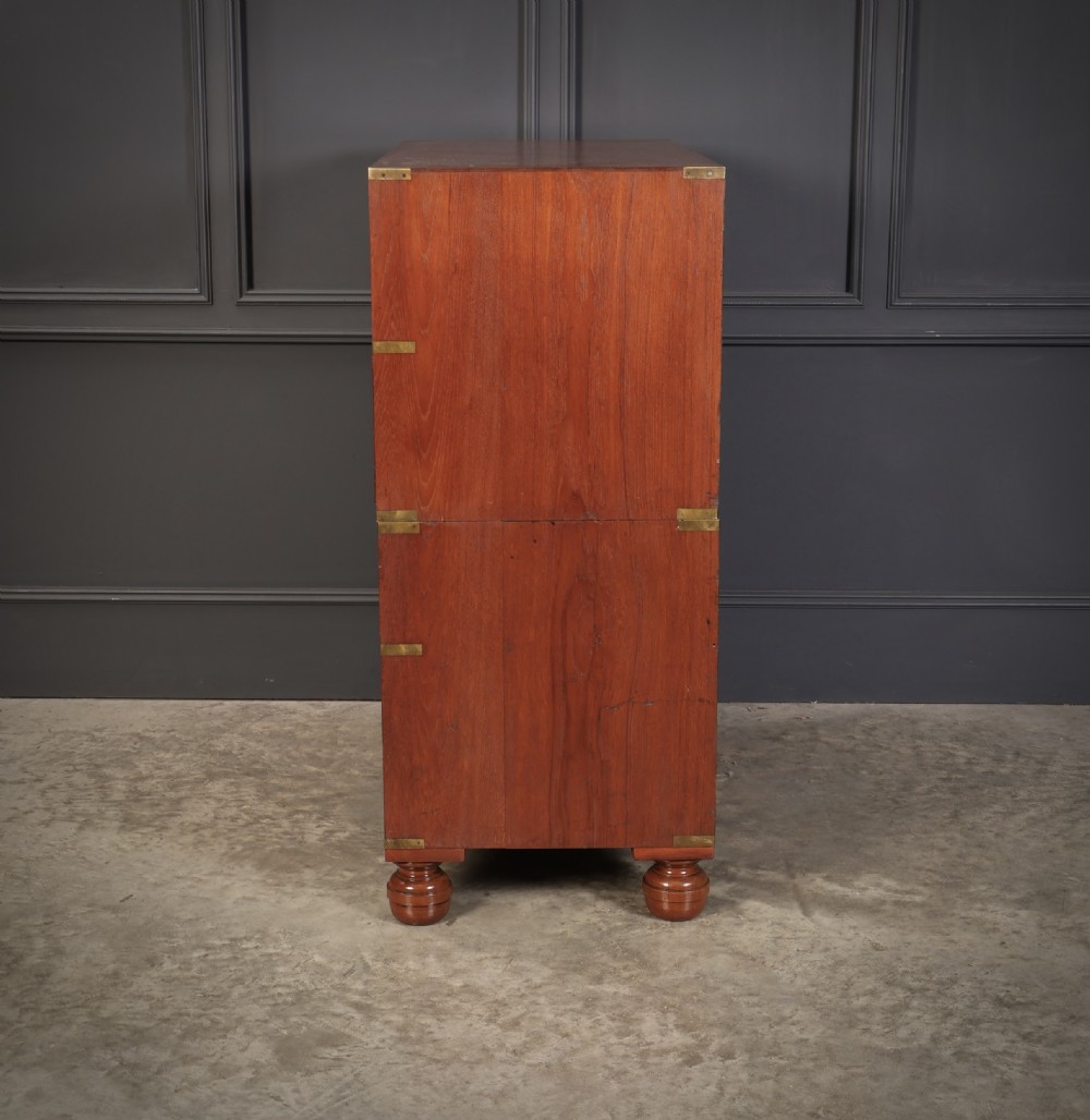 A Victorian teak military campaign secretaire chest featuring an open fall-front desk compartment, flush brass handles, brass corner brackets, and turned feet against a dark grey panelled wall. View X
