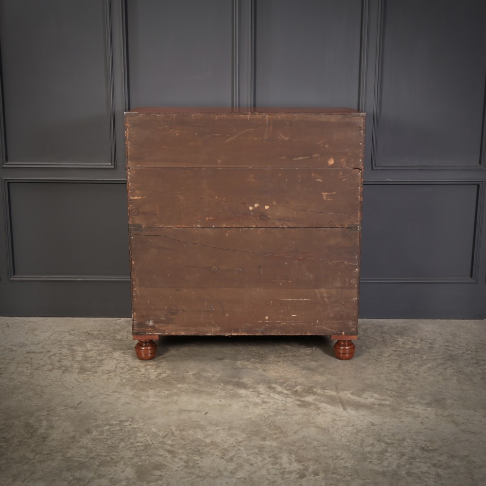 A Victorian teak military campaign secretaire chest featuring an open fall-front desk compartment, flush brass handles, brass corner brackets, and turned feet against a dark grey panelled wall. View XI