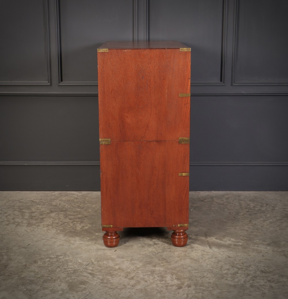 A Victorian teak military campaign secretaire chest featuring an open fall-front desk compartment, flush brass handles, brass corner brackets, and turned feet against a dark grey panelled wall. View XII
