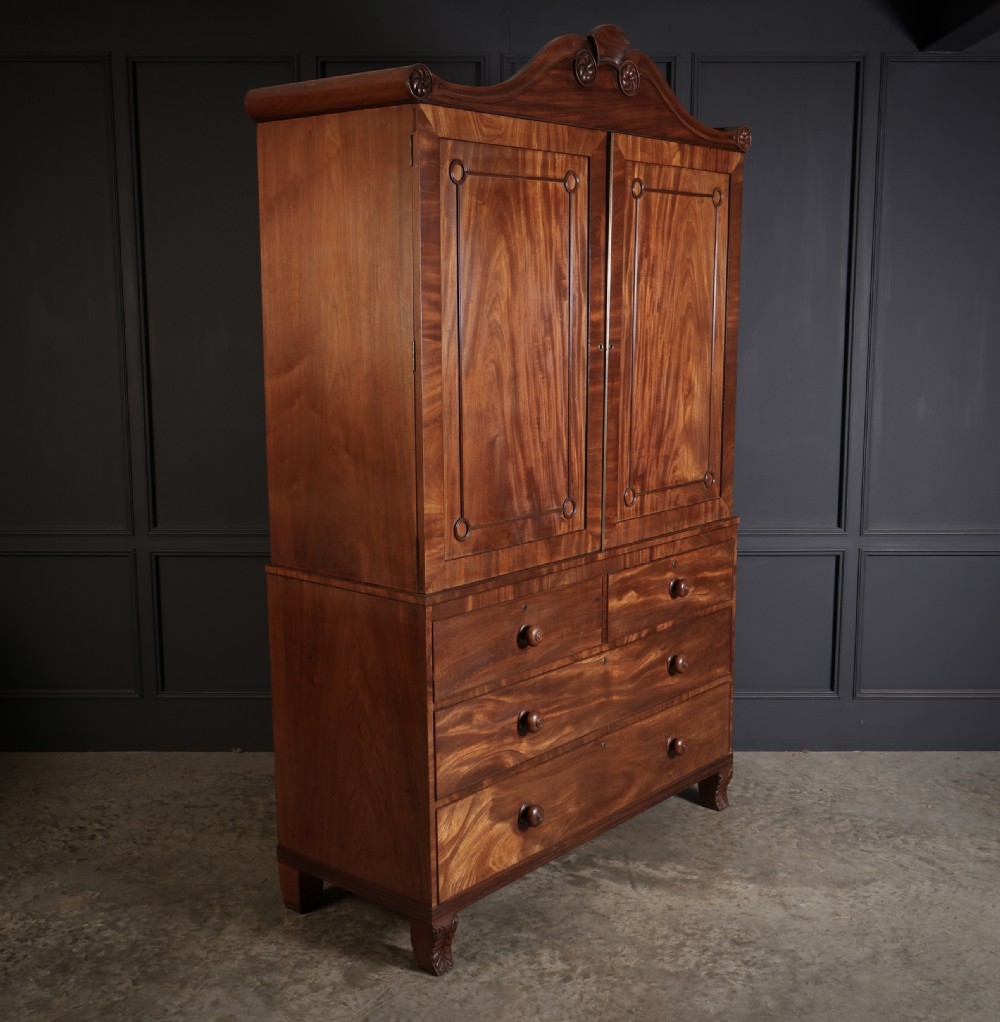 A late Regency mahogany linen press featuring a carved scrolled pediment, double panelled doors with flame mahogany veneers, and a base with four drawers. View III