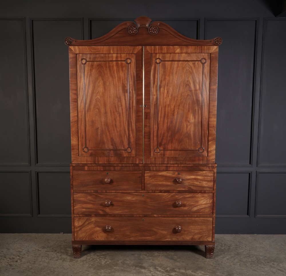 A late Regency mahogany linen press featuring a carved scrolled pediment, double panelled doors with flame mahogany veneers, and a base with four drawers.