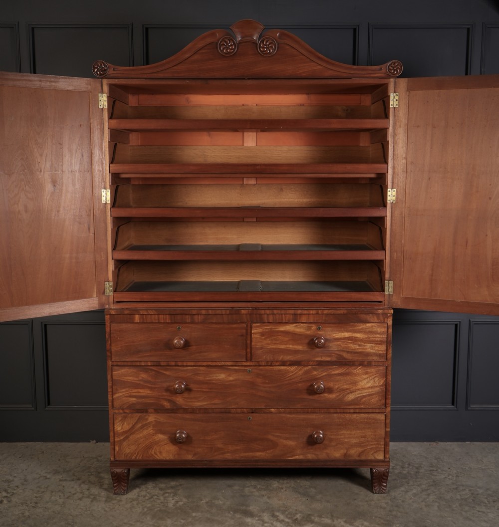 A late Regency mahogany linen press featuring a carved scrolled pediment, double panelled doors with flame mahogany veneers, and a base with four drawers. View V