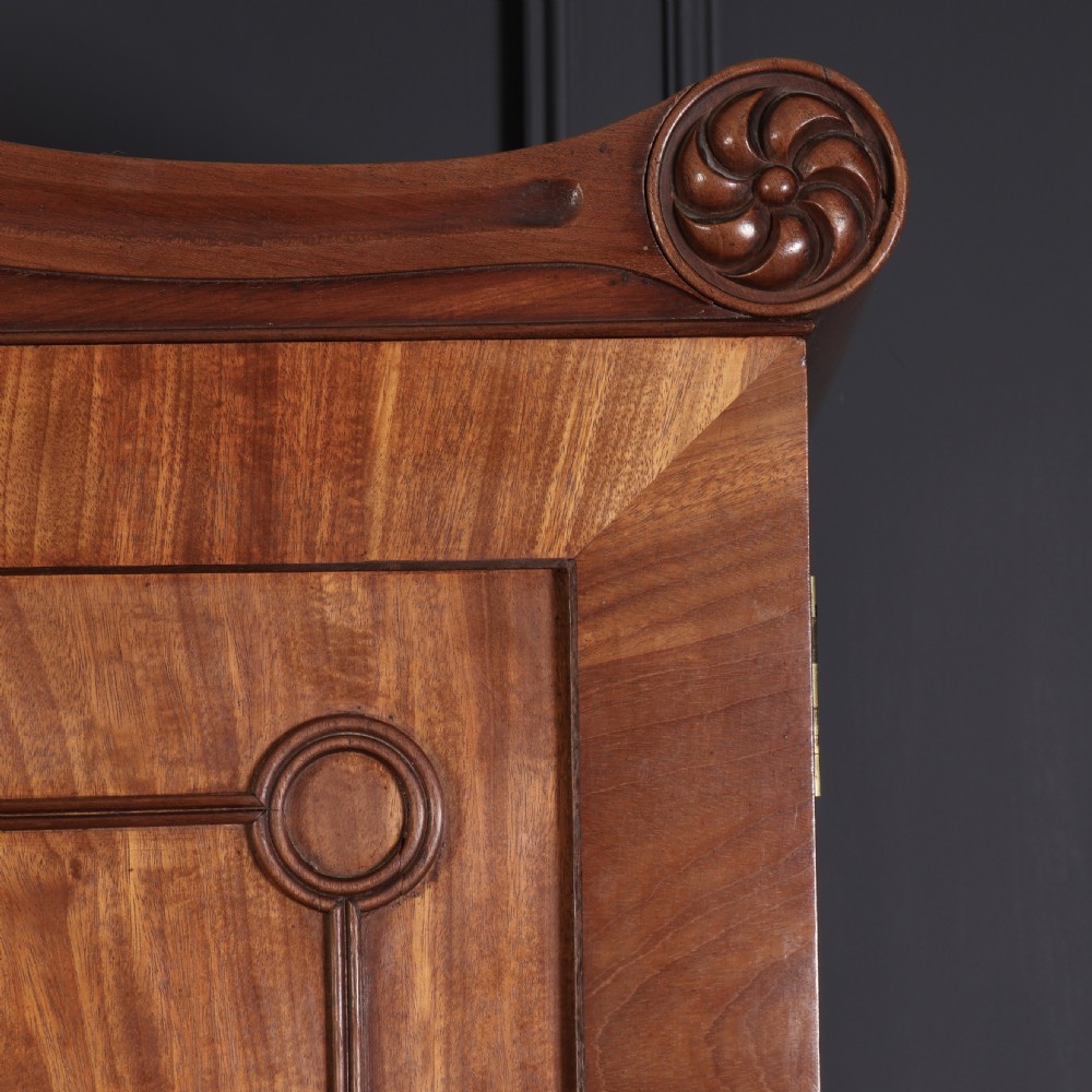 A late Regency mahogany linen press featuring a carved scrolled pediment, double panelled doors with flame mahogany veneers, and a base with four drawers. View VII