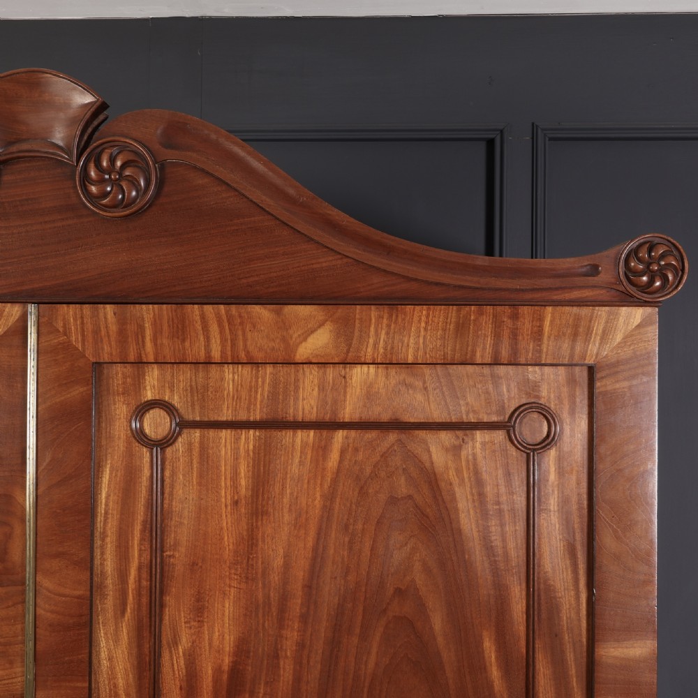 A late Regency mahogany linen press featuring a carved scrolled pediment, double panelled doors with flame mahogany veneers, and a base with four drawers. View VIII