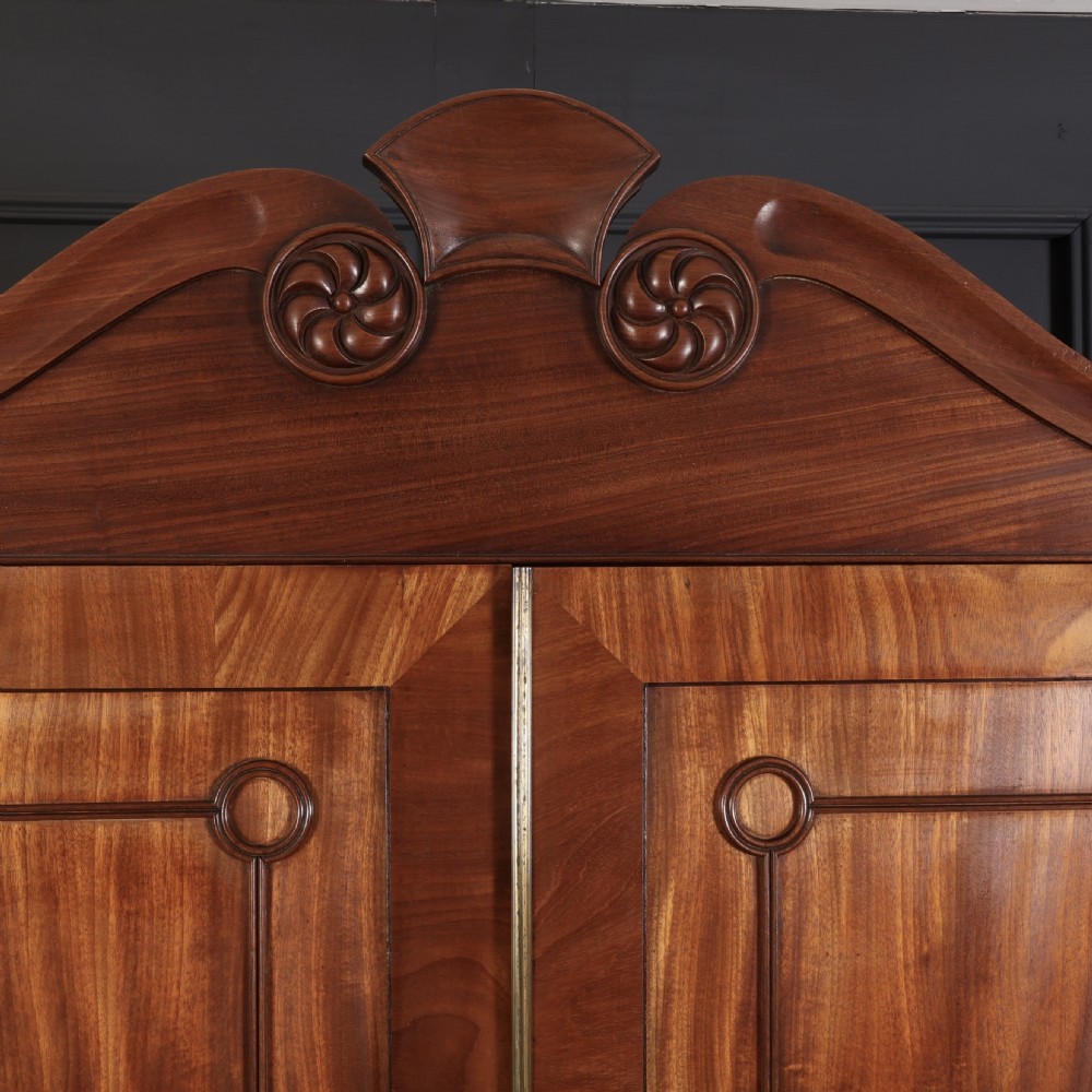 A late Regency mahogany linen press featuring a carved scrolled pediment, double panelled doors with flame mahogany veneers, and a base with four drawers. View IX