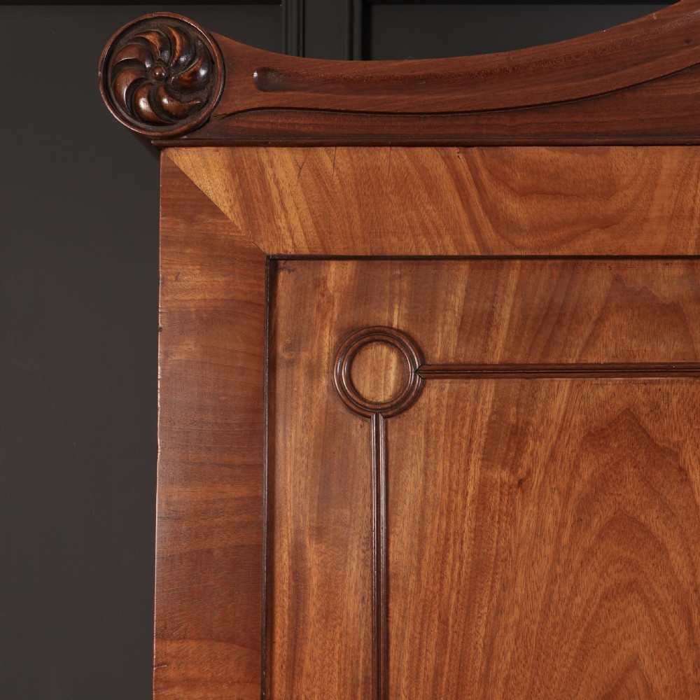 A late Regency mahogany linen press featuring a carved scrolled pediment, double panelled doors with flame mahogany veneers, and a base with four drawers. View XI