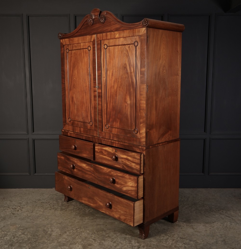 A late Regency mahogany linen press featuring a carved scrolled pediment, double panelled doors with flame mahogany veneers, and a base with four drawers. View XIII