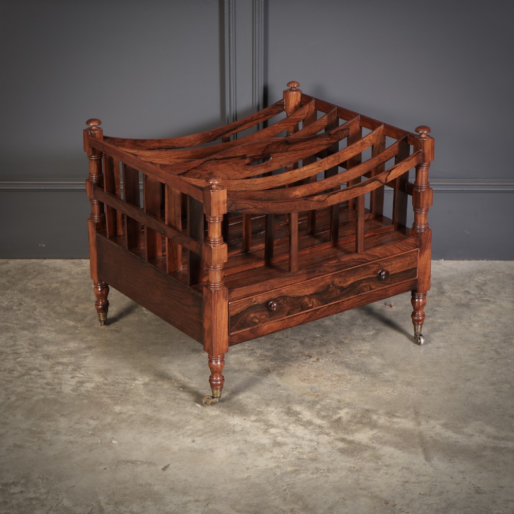 A Regency dark rosewood Canterbury music rack featuring concave slatted dividers, turned corner posts with finials, a lower drawer, and brass castors.