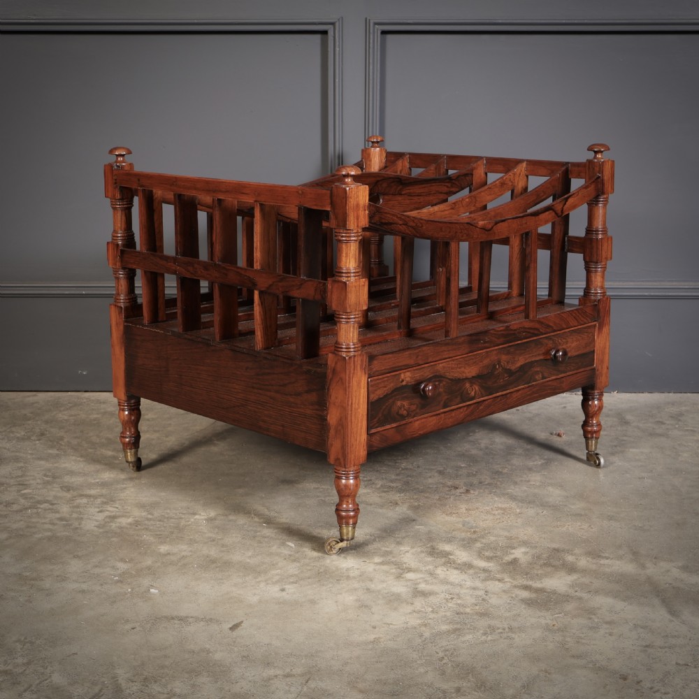 A Regency dark rosewood Canterbury music rack featuring concave slatted dividers, turned corner posts with finials, a lower drawer, and brass castors. View II