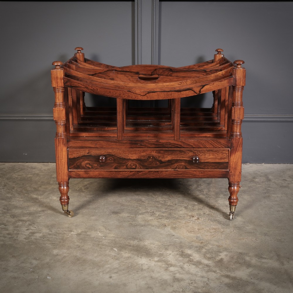 A Regency dark rosewood Canterbury music rack featuring concave slatted dividers, turned corner posts with finials, a lower drawer, and brass castors. View IV