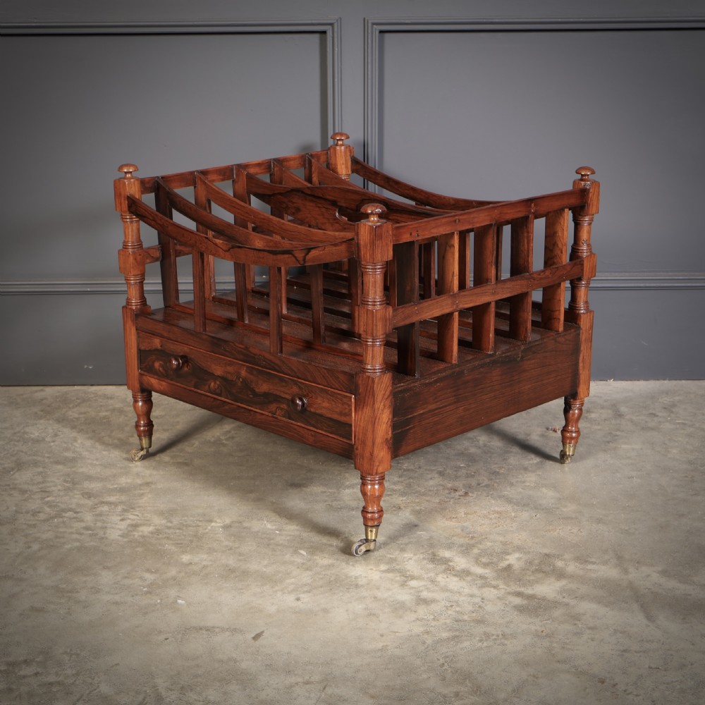 A Regency dark rosewood Canterbury music rack featuring concave slatted dividers, turned corner posts with finials, a lower drawer, and brass castors. View V