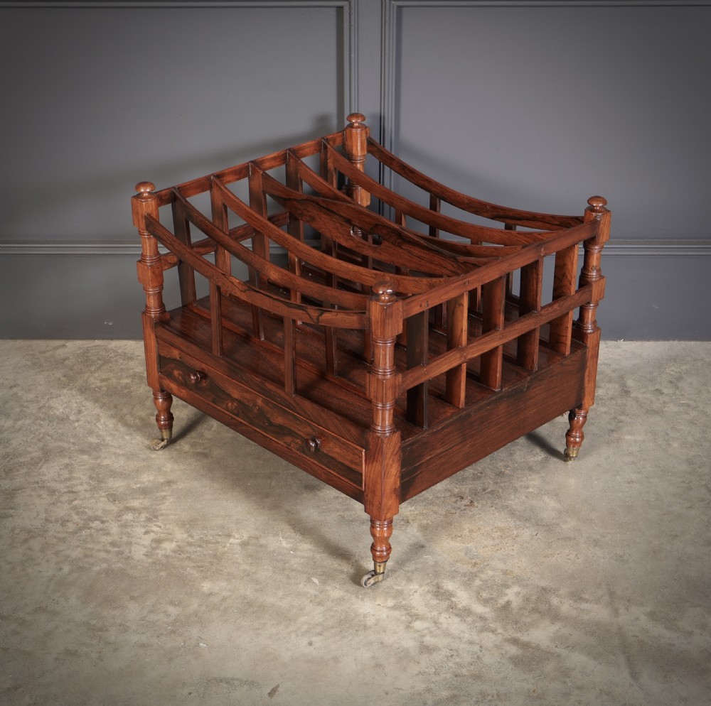 A Regency dark rosewood Canterbury music rack featuring concave slatted dividers, turned corner posts with finials, a lower drawer, and brass castors. View VI