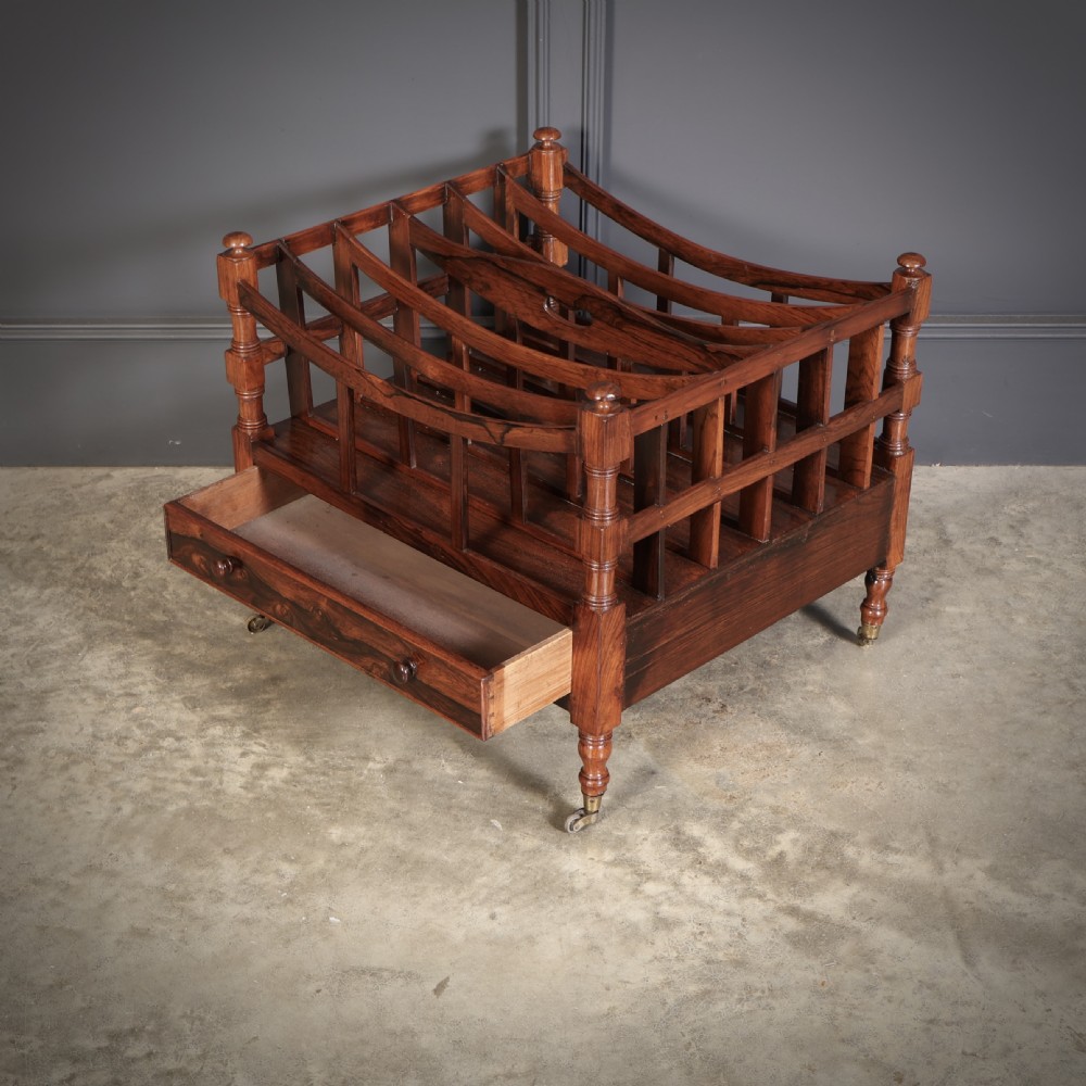 A Regency dark rosewood Canterbury music rack featuring concave slatted dividers, turned corner posts with finials, a lower drawer, and brass castors. View VII