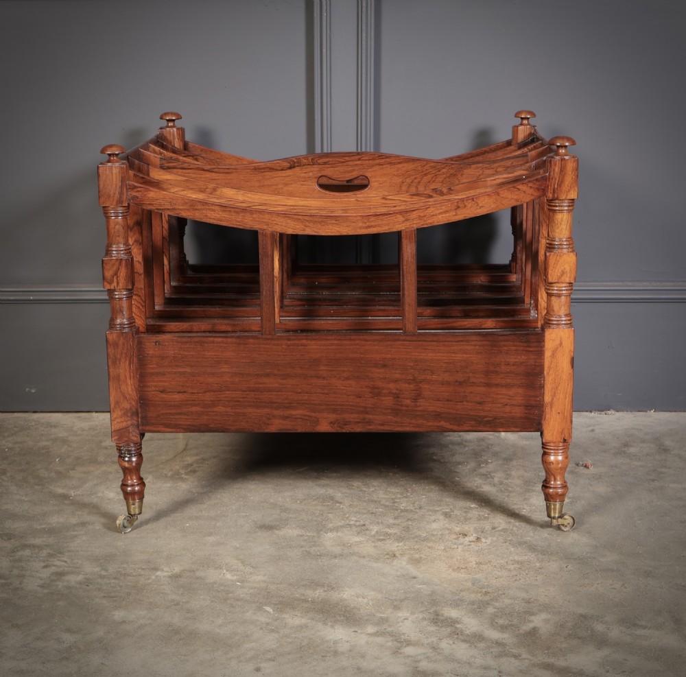 A Regency dark rosewood Canterbury music rack featuring concave slatted dividers, turned corner posts with finials, a lower drawer, and brass castors. View IX