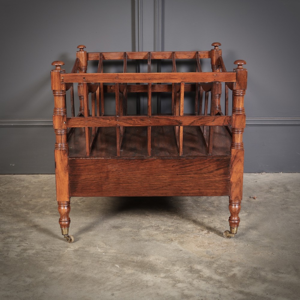 A Regency dark rosewood Canterbury music rack featuring concave slatted dividers, turned corner posts with finials, a lower drawer, and brass castors. View X