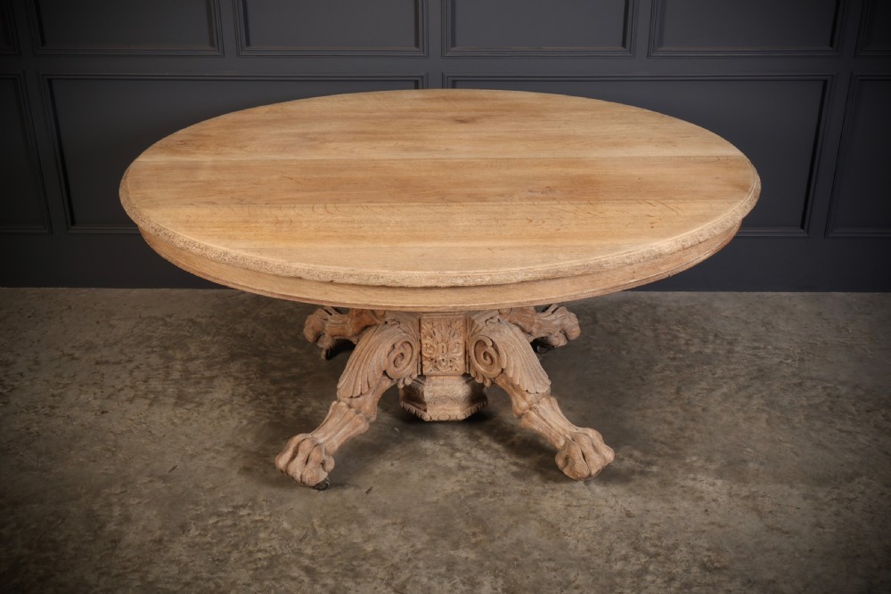 An oval bleached oak centre table featuring an intricately carved pedestal base with scroll legs and paw feet.