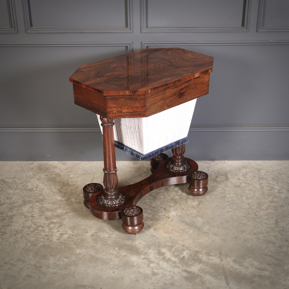 A William IV rosewood sewing table featuring an octagonal hinged top, a pleated fabric work basket, and carved twin column supports on a plinth base.
