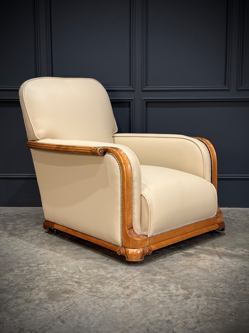 A pair of Art Deco armchairs featuring cream leather upholstery, polished walnut arm supports with scroll details, and metal studwork trim. View V