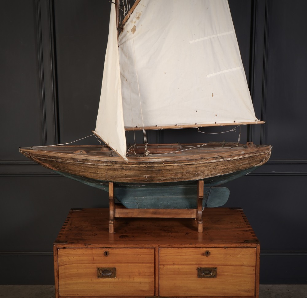 An antique wooden pond yacht with off-white fabric sails, a carved brown hull with a teal painted lower section, mounted on a wooden display stand. View III