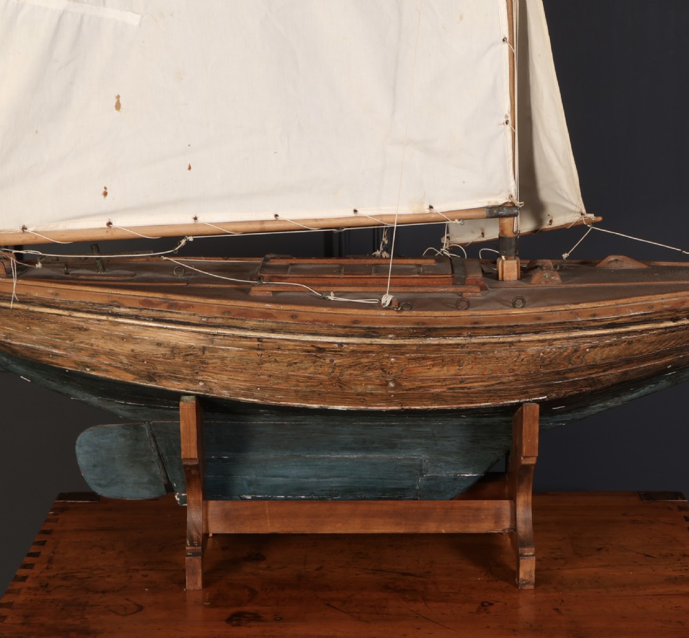 An antique wooden pond yacht with off-white fabric sails, a carved brown hull with a teal painted lower section, mounted on a wooden display stand. View II