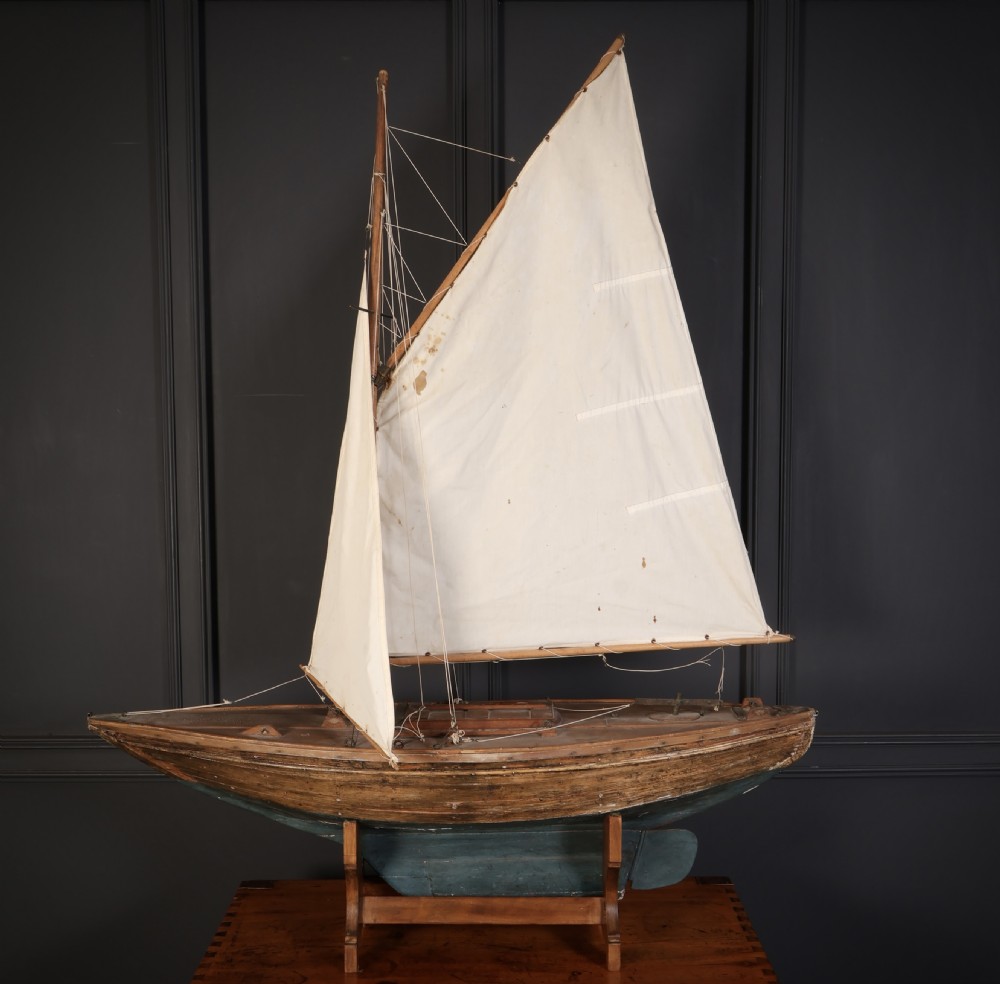 An antique wooden pond yacht with off-white fabric sails, a carved brown hull with a teal painted lower section, mounted on a wooden display stand.