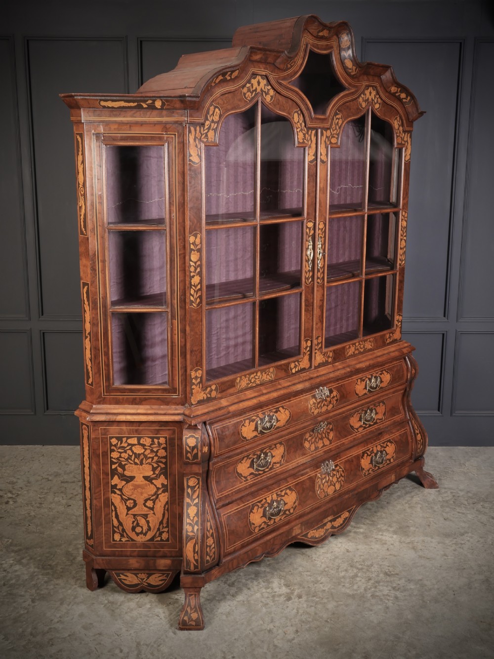 A Dutch Baroque style walnut display cabinet featuring floral marquetry, a glazed double-door upper section, and a three-drawer bombe base.