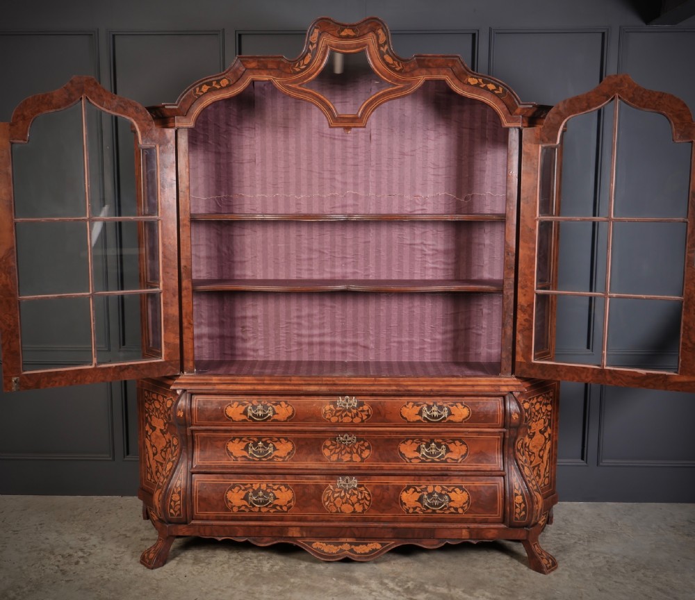A Dutch Baroque style walnut display cabinet featuring floral marquetry, a glazed double-door upper section, and a three-drawer bombe base. View VI