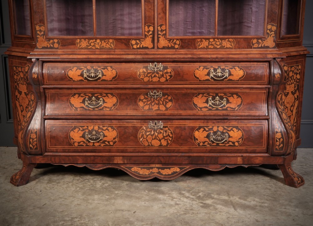 A Dutch Baroque style walnut display cabinet featuring floral marquetry, a glazed double-door upper section, and a three-drawer bombe base. View VII