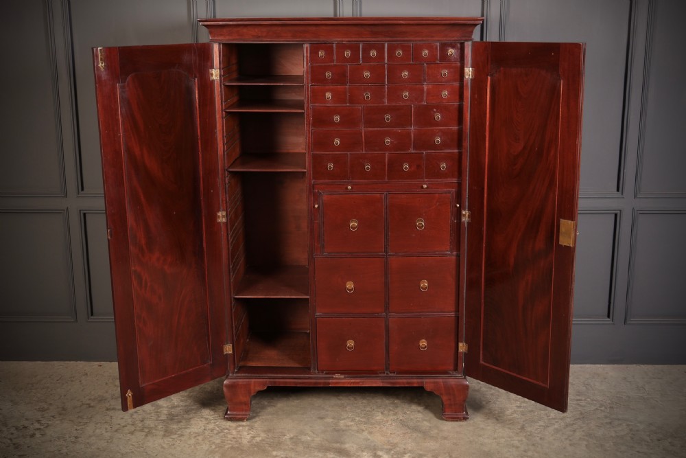 A George III mahogany estate cabinet shown with open panelled doors, revealing an interior with shelves on one side and multiple fitted drawers with brass ring handles on the other.