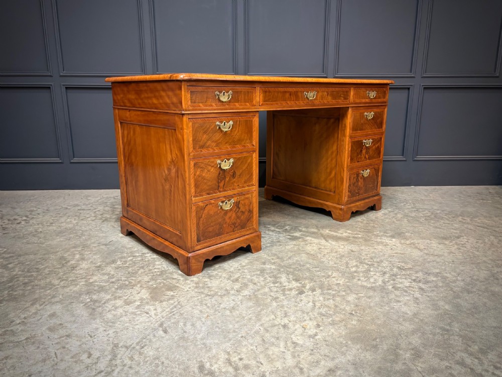 An antique burr walnut pedestal desk featuring inlaid feather-banded borders, brass drop handles, and multiple side drawers. View VI