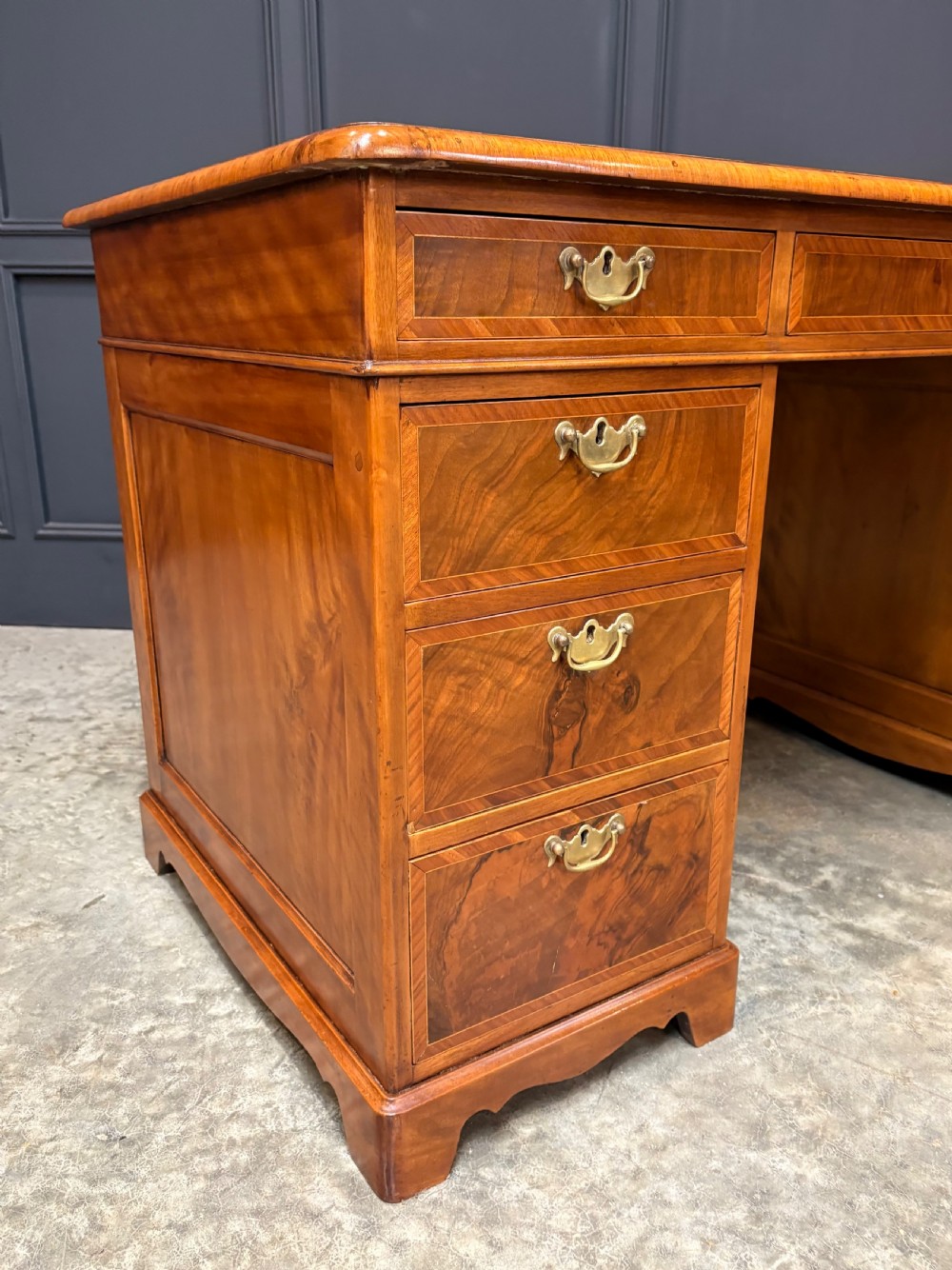 An antique burr walnut pedestal desk featuring inlaid feather-banded borders, brass drop handles, and multiple side drawers. View VII