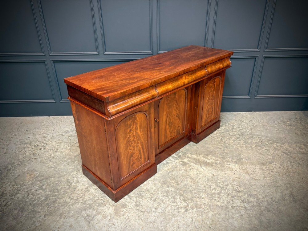 A Victorian mahogany sideboard featuring three flame mahogany veneer arched panel doors and concealed frieze drawers set on a plinth base. View VI