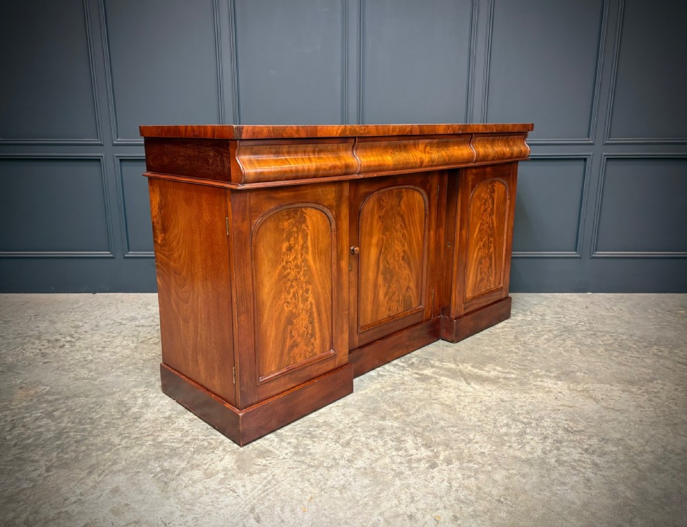 A Victorian mahogany sideboard featuring three flame mahogany veneer arched panel doors and concealed frieze drawers set on a plinth base. View II