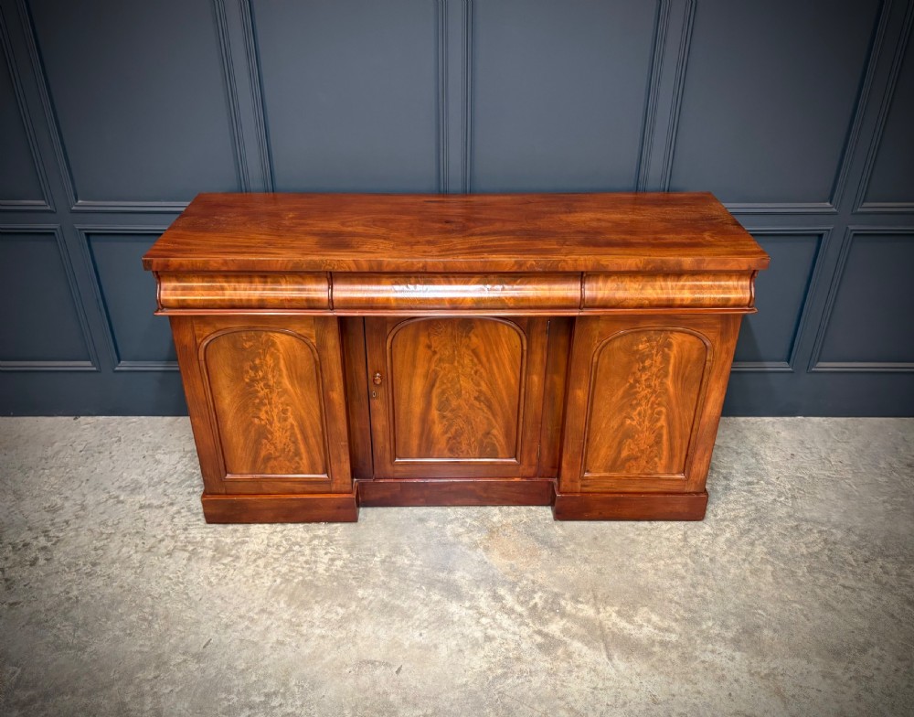 A Victorian mahogany sideboard featuring three flame mahogany veneer arched panel doors and concealed frieze drawers set on a plinth base.