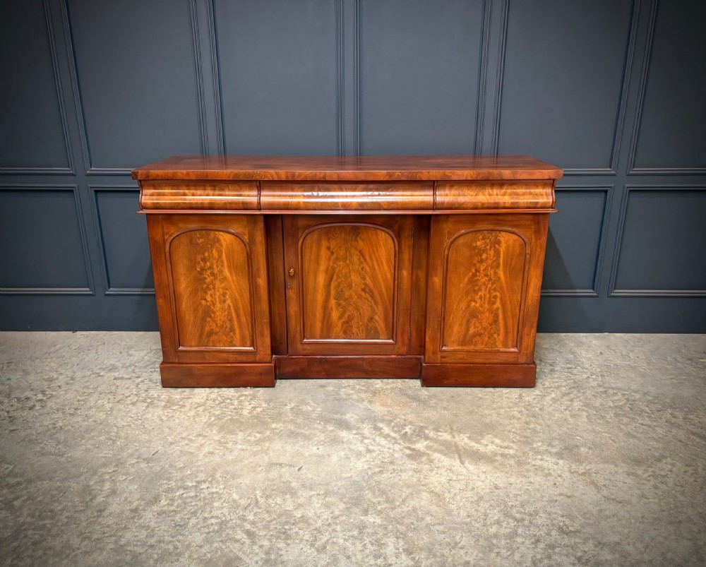A Victorian mahogany sideboard featuring three flame mahogany veneer arched panel doors and concealed frieze drawers set on a plinth base. View V