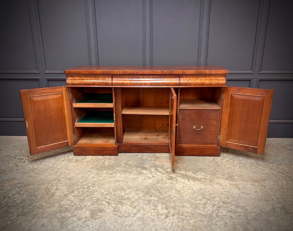 A Victorian mahogany sideboard featuring three flame mahogany veneer arched panel doors and concealed frieze drawers set on a plinth base. View VII