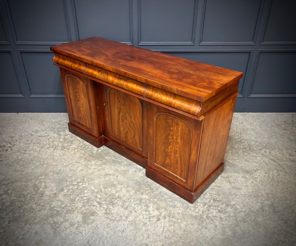 A Victorian mahogany sideboard featuring three flame mahogany veneer arched panel doors and concealed frieze drawers set on a plinth base. View VIII