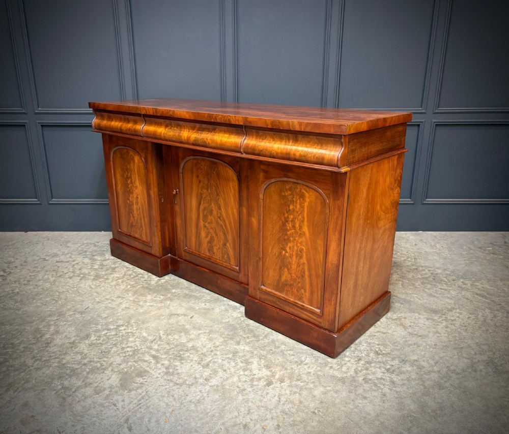 A Victorian mahogany sideboard featuring three flame mahogany veneer arched panel doors and concealed frieze drawers set on a plinth base. View IX