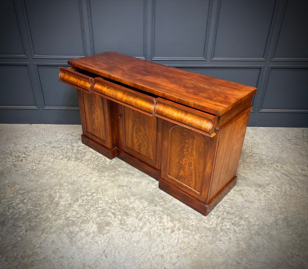 A Victorian mahogany sideboard featuring three flame mahogany veneer arched panel doors and concealed frieze drawers set on a plinth base. View X
