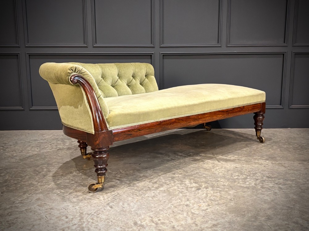 A Victorian rosewood chaise longue upholstered in olive green velvet with deep buttoning along the asymmetrical backrest, supported by turned legs on brass castors. View III