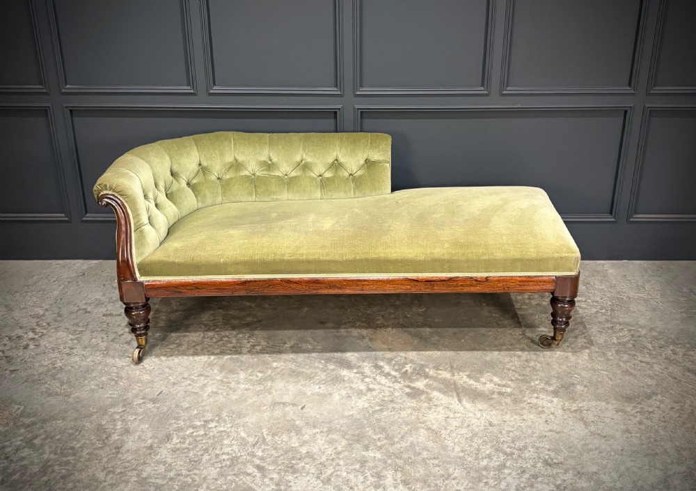 A Victorian rosewood chaise longue upholstered in olive green velvet with deep buttoning along the asymmetrical backrest, supported by turned legs on brass castors. View II