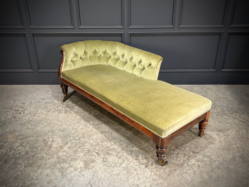 A Victorian rosewood chaise longue upholstered in olive green velvet with deep buttoning along the asymmetrical backrest, supported by turned legs on brass castors. View V