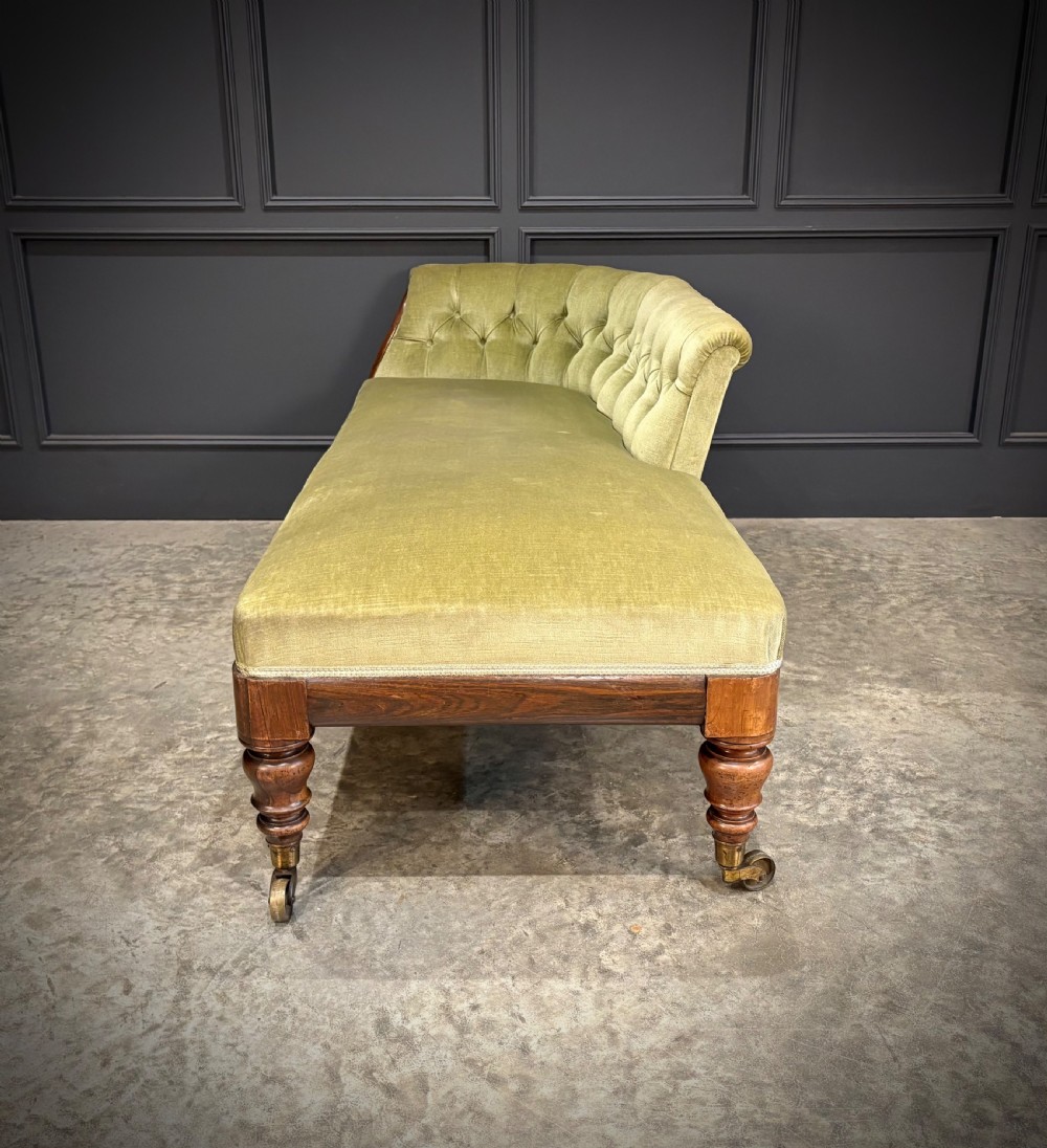 A Victorian rosewood chaise longue upholstered in olive green velvet with deep buttoning along the asymmetrical backrest, supported by turned legs on brass castors. View VII