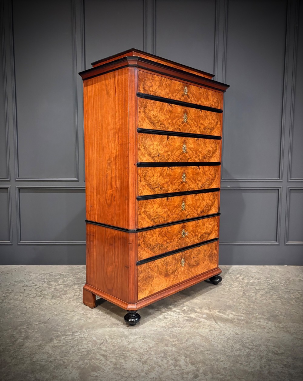 A tall 19th-century burr walnut chest of drawers featuring seven graduated drawers with brass escutcheons and ebonised mouldings set on turned front feet.